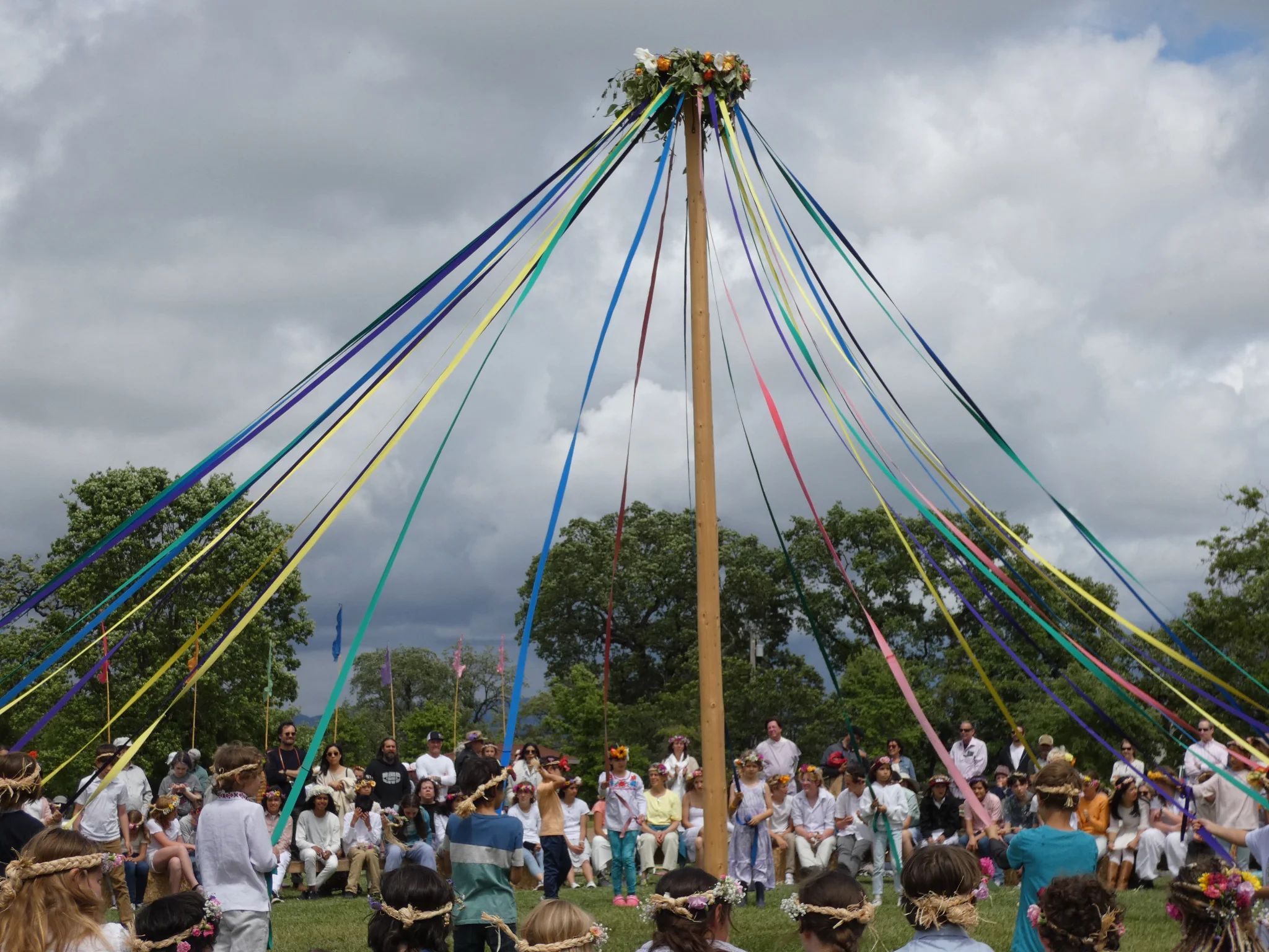 May Faire pole with streamers and dancers and audience, cloudy sky in background