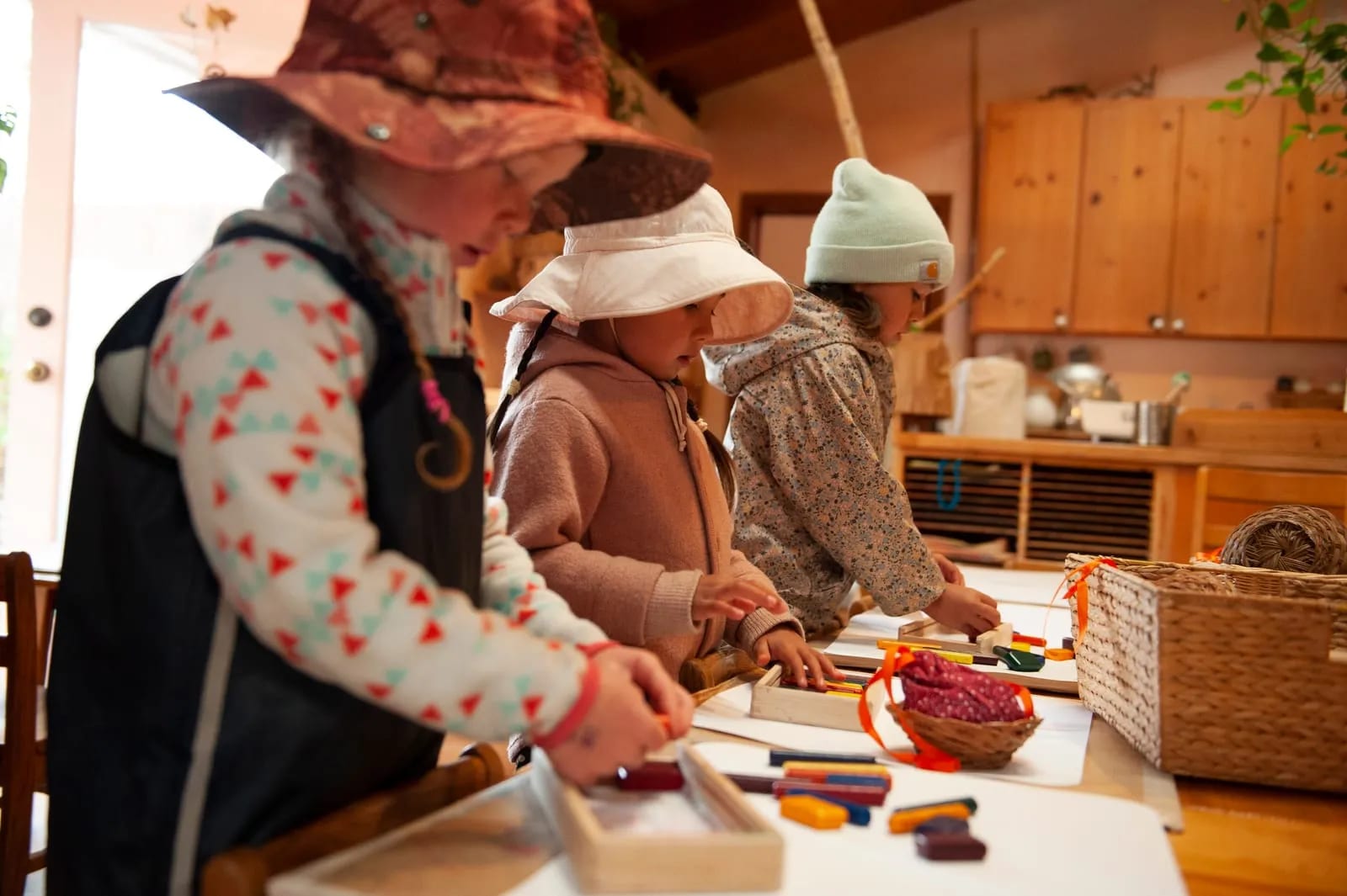 Three young children in sun hats stand at a table making wax-crayon block prints, watched over in a warm wood-paneled classroom