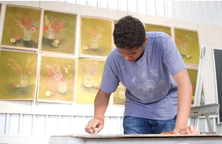 A student leans over a worktable in front of a wall of pastel still-life paintings of flowers in jars, part of a Grade 8 art project