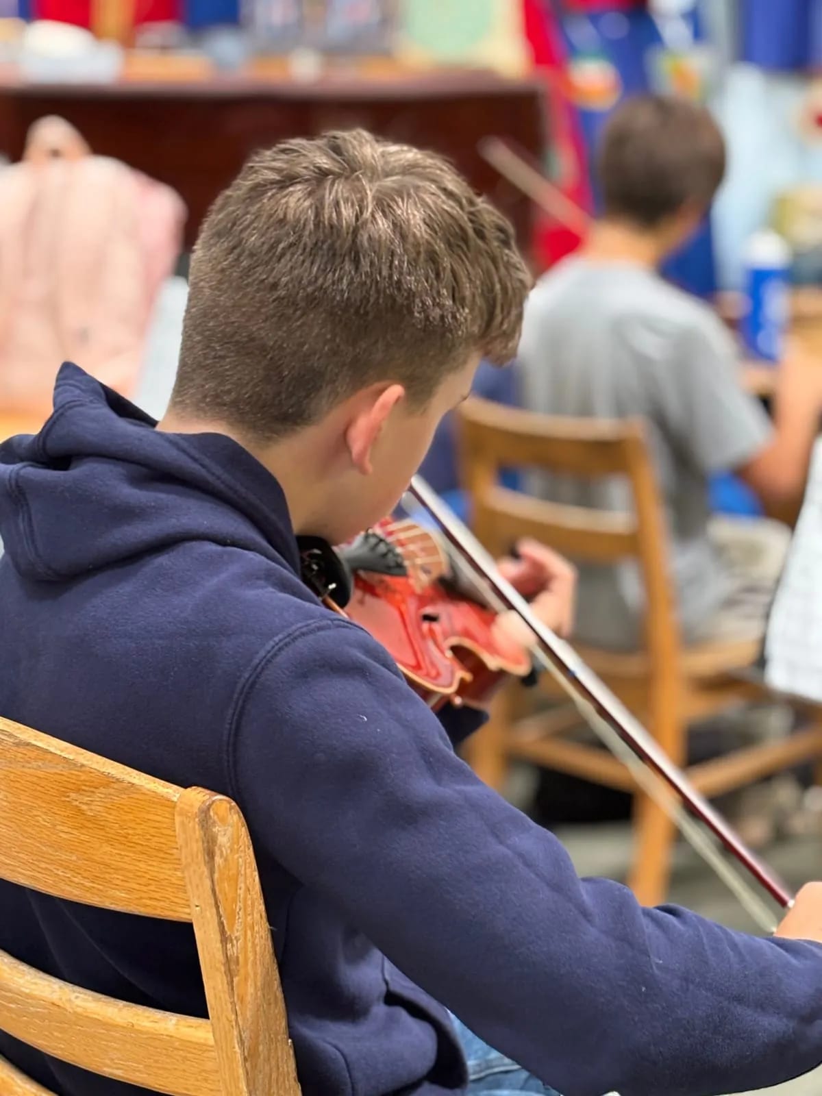 A student seen from behind draws a bow across a violin during a Lower School music class