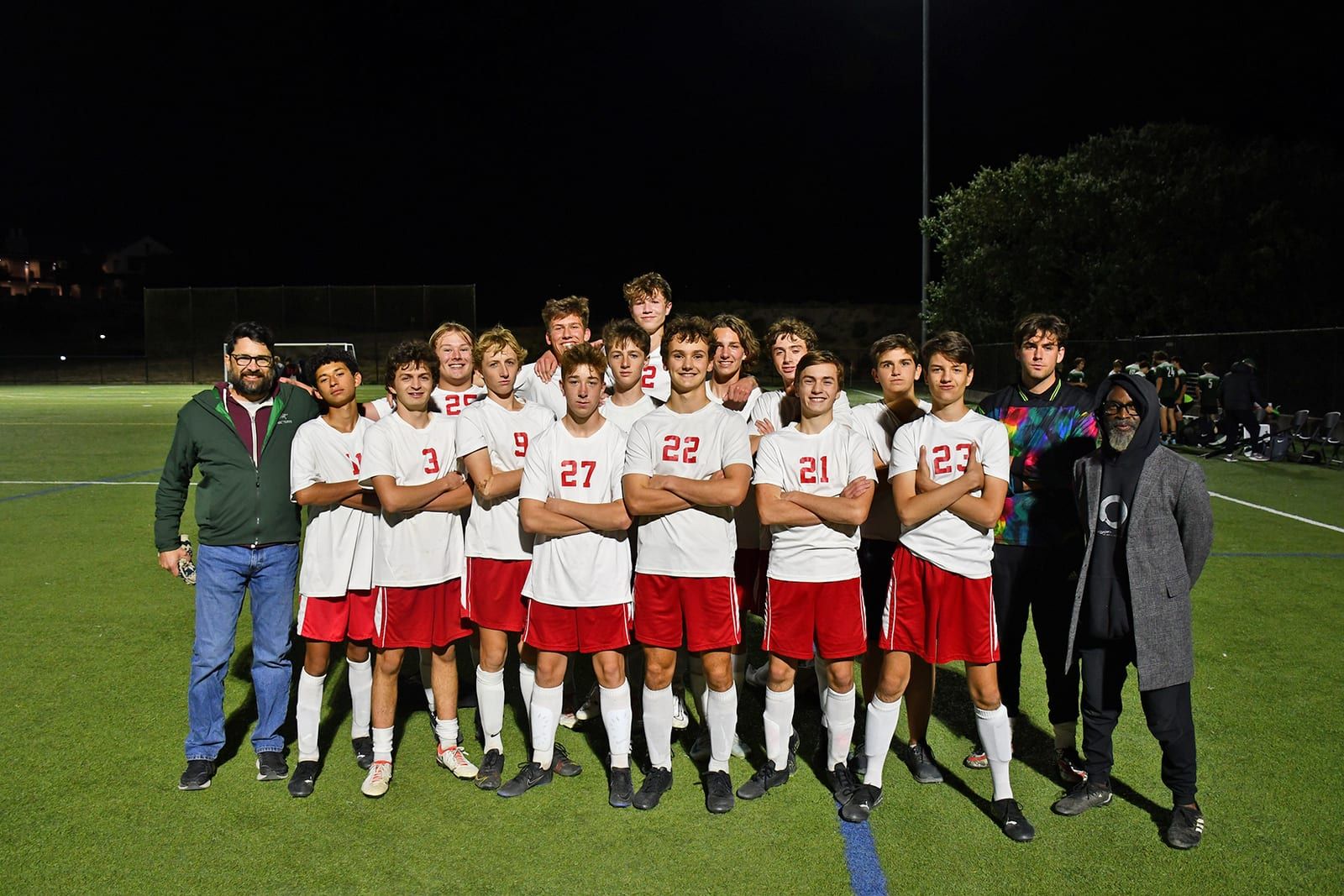 The high school boys soccer team poses together in white and red uniforms on a turf field at night under stadium lights, flanked by two coaches.
