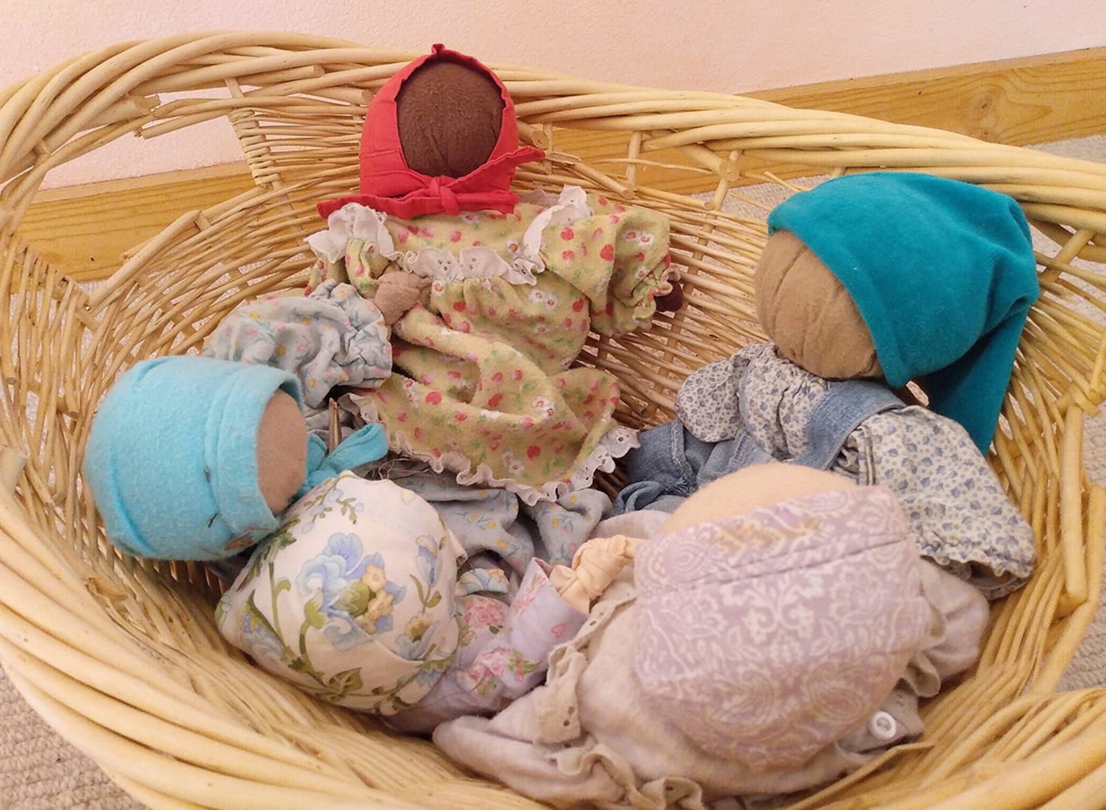 A woven wicker basket cradles five soft handmade Waldorf dolls, each with a simple stitched face and a different colored knit bonnet, tucked together as if napping.