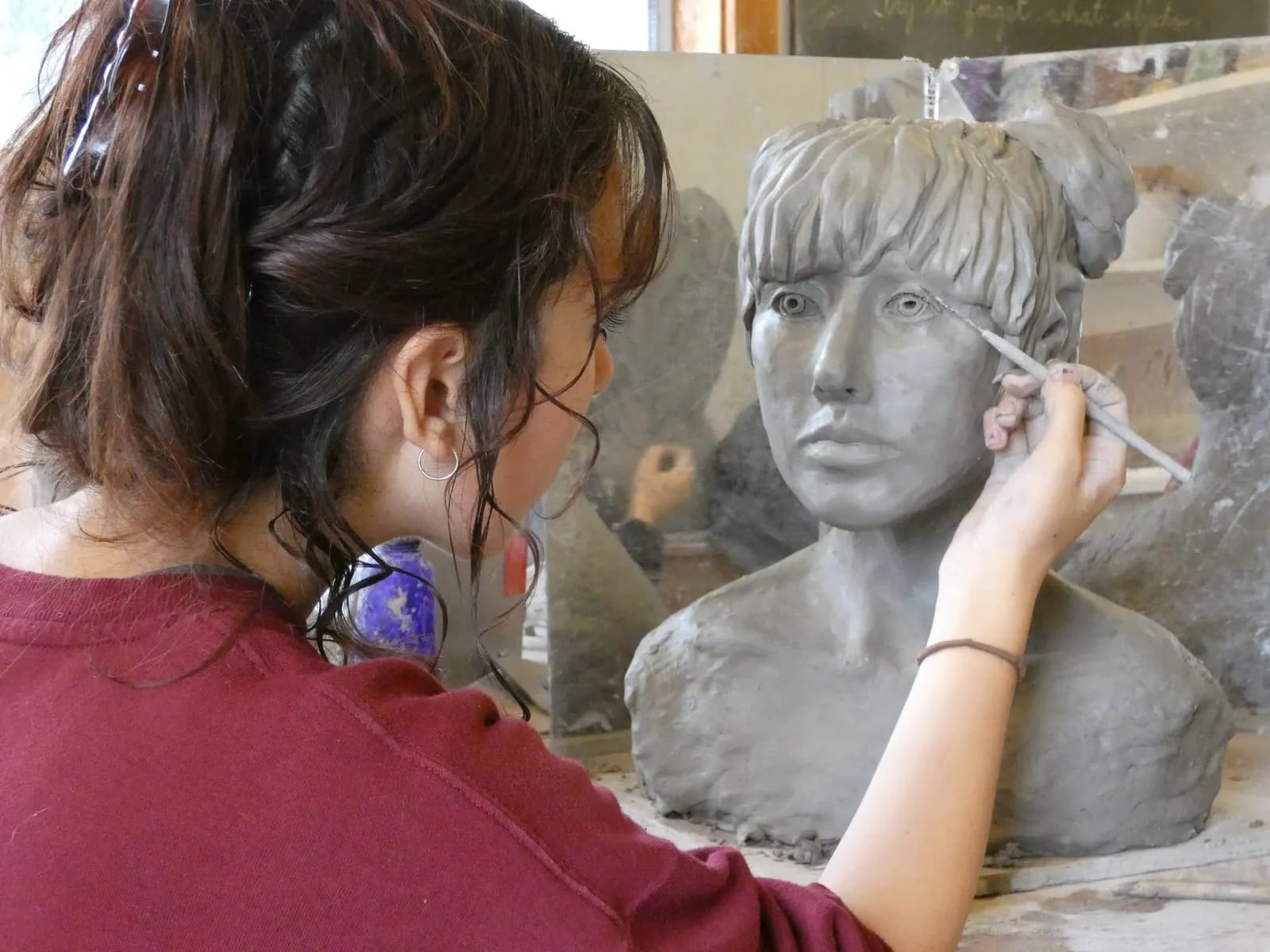 A student models a life-size portrait bust in clay, working from a mirror.