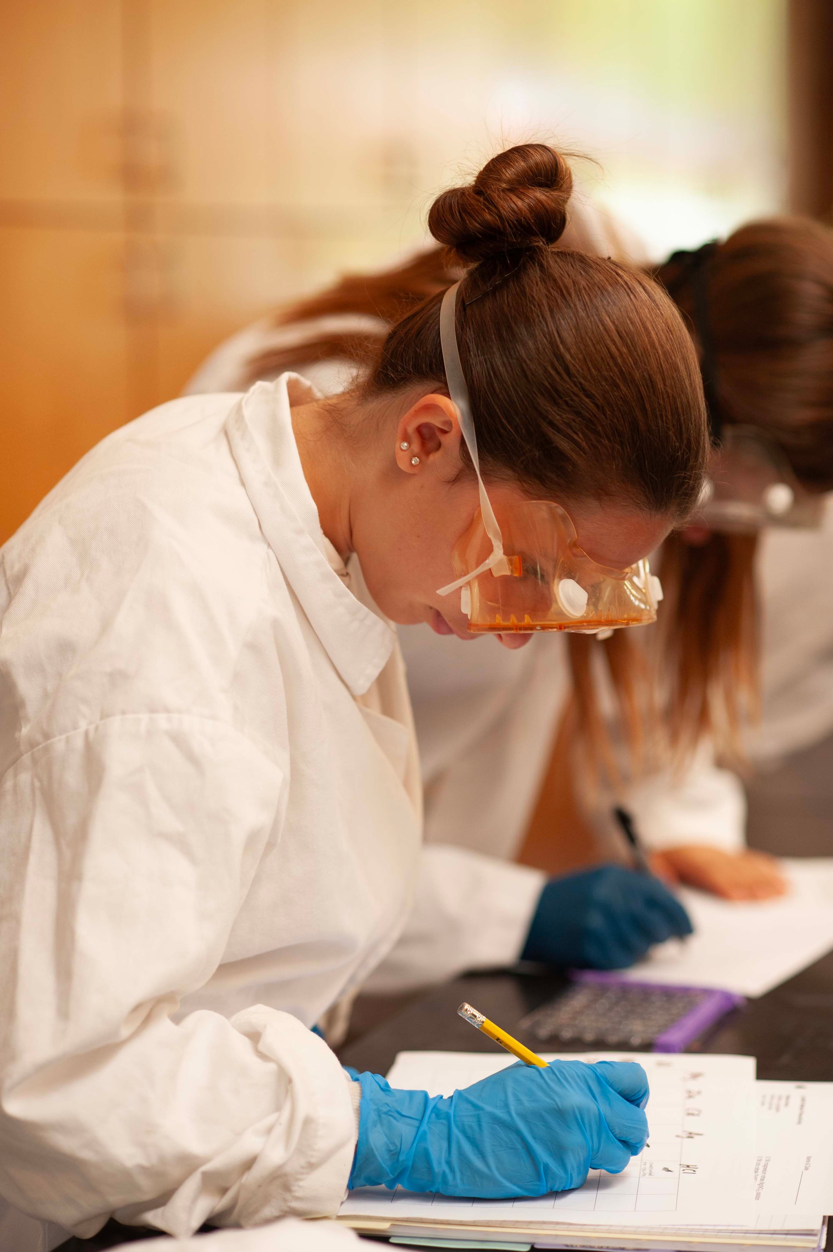 A high school student in a white lab coat, safety goggles and blue nitrile gloves bends over her lab notebook, pencil in hand, recording a result; a classmate works at the bench behind her.