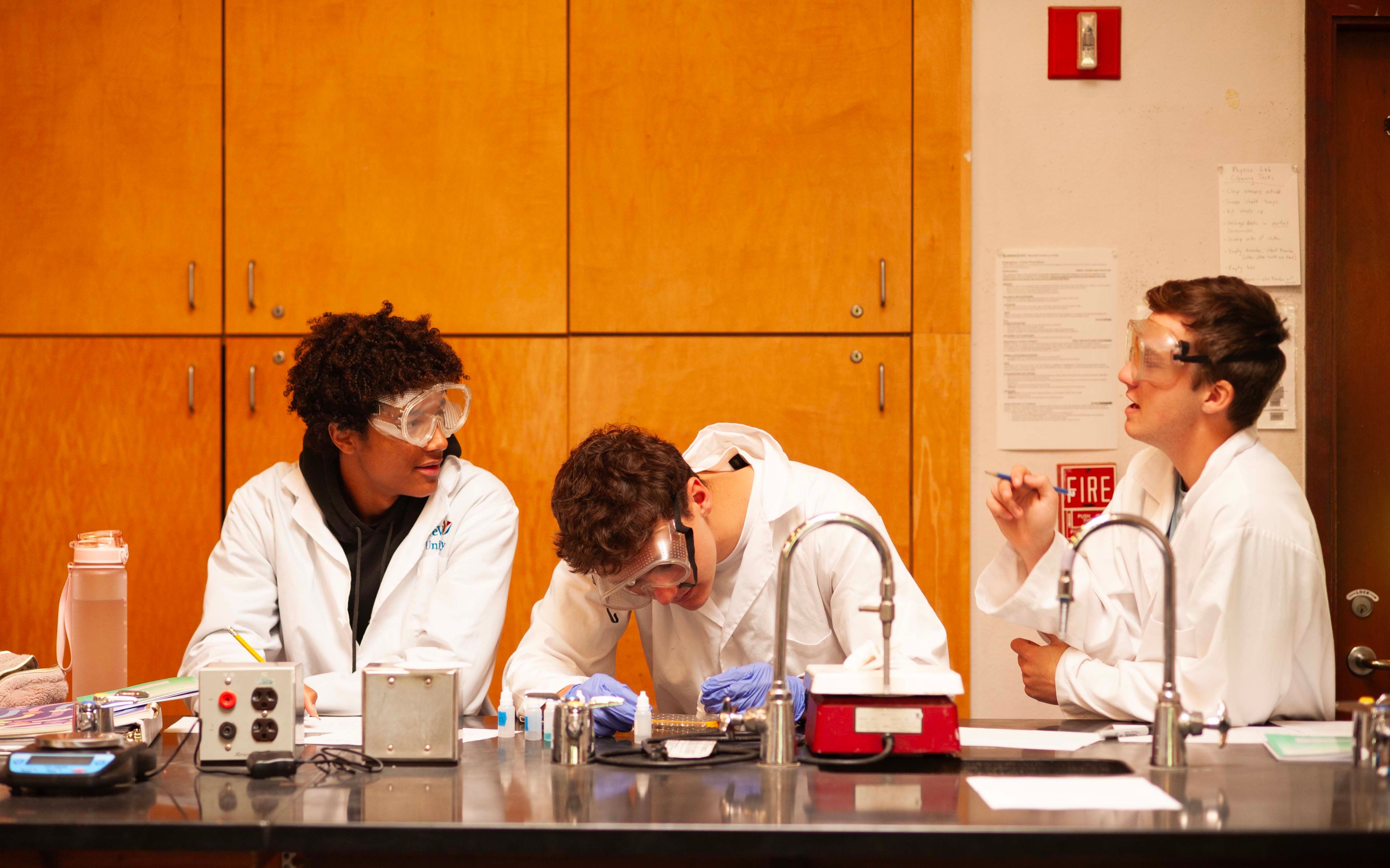 Three High School students in lab coats and safety goggles working together at a science bench