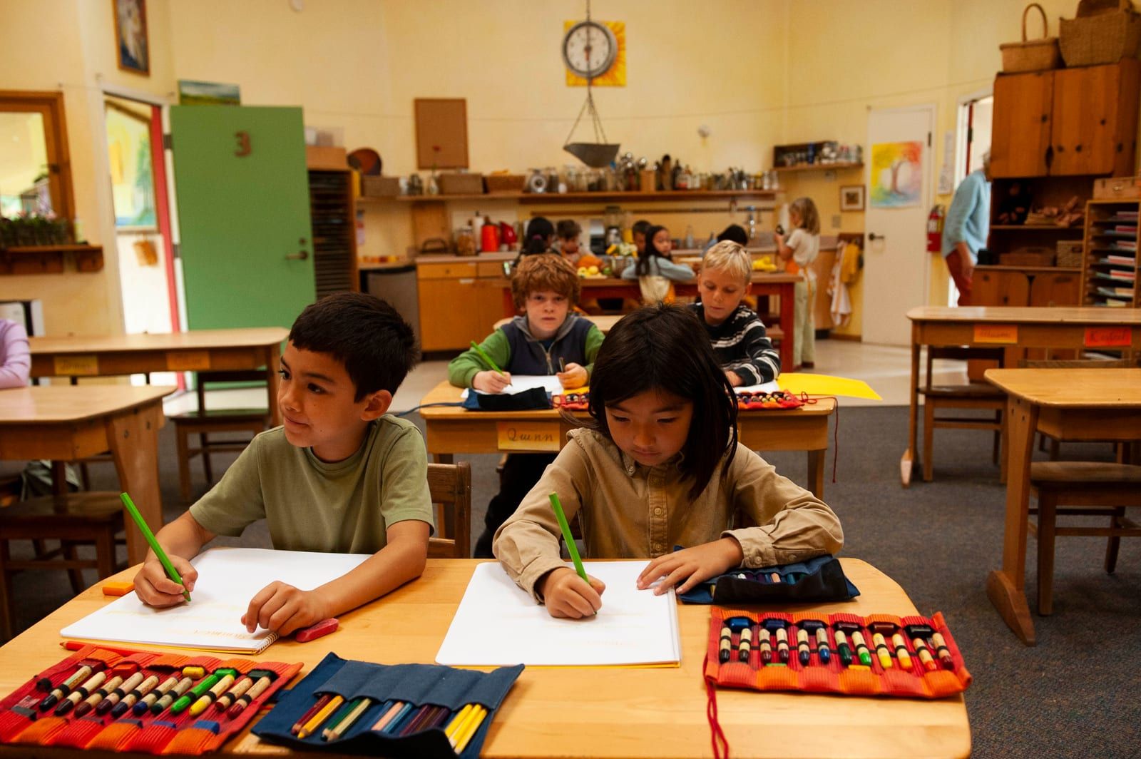 Two lower-school students sit at a wooden desk writing with colored pencils in a sunlit classroom, with more students and open art-supply rolls visible around the room
