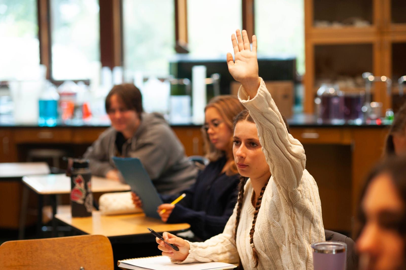 A high school student in a cream sweater raises her hand to answer a question in a science classroom, with classmates seated at lab tables in front of her