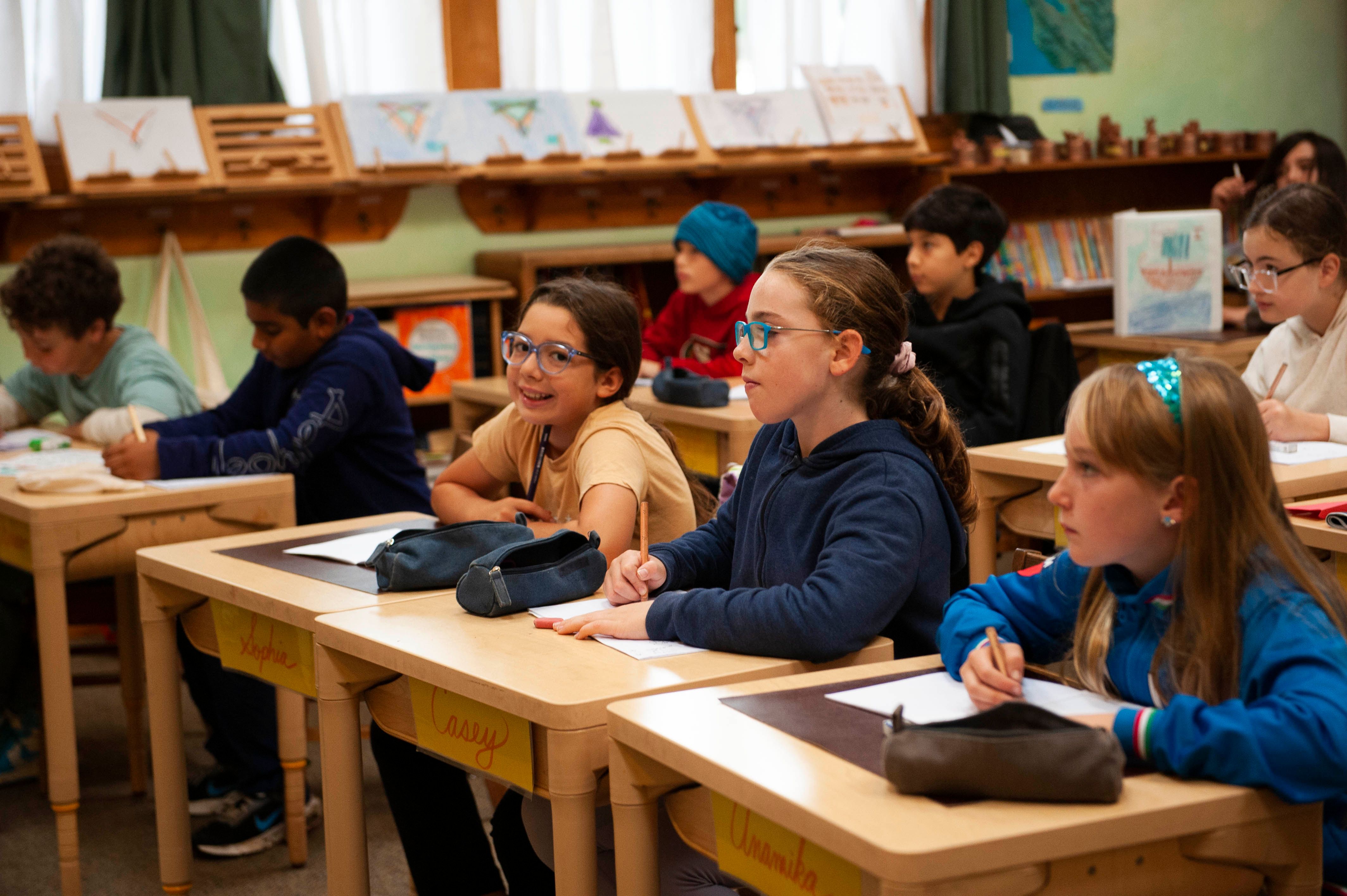 Lower-school students sit at wooden desks with pencils and main-lesson books, looking toward the front of a green-walled classroom, their geometry drawings propped along a shelf rail behind them and hand-lettered name cards on the desk fronts