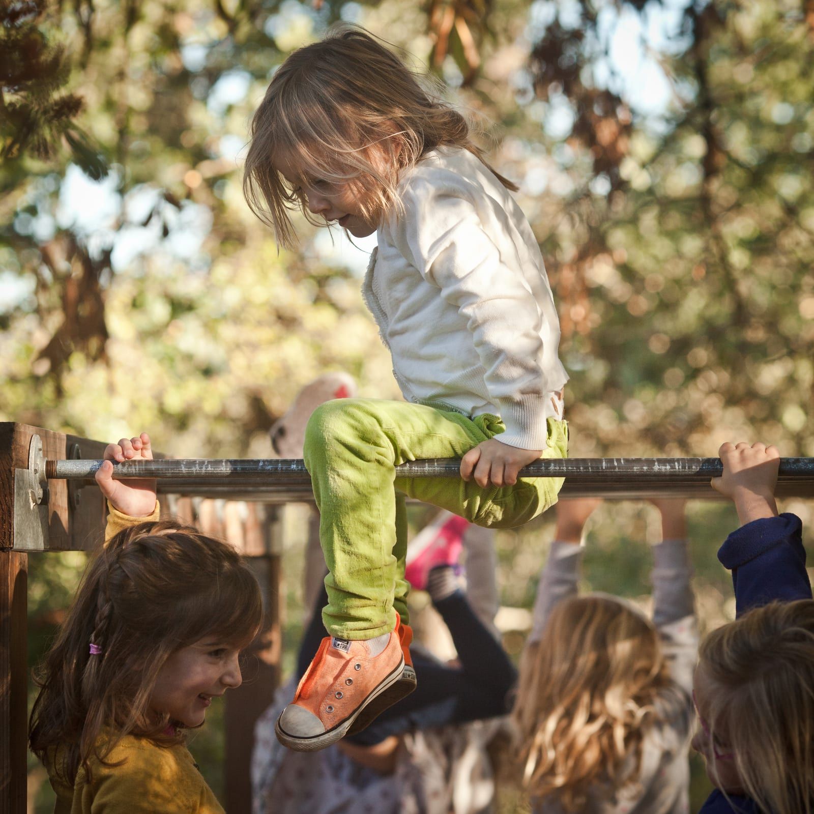 A preschooler in green pants and orange sneakers balances astride a wooden monkey-bar rail outdoors, other children hanging beneath the bar in dappled forest light.