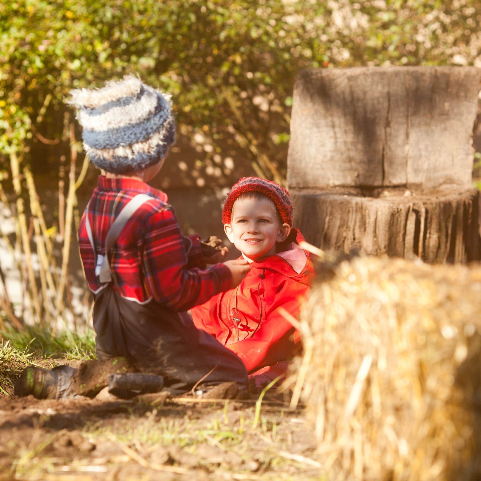 Two small children in red and plaid outerwear sit together on the ground near a hay bale and tree stump, one smiling at the camera while the other reaches toward him with muddy hands.