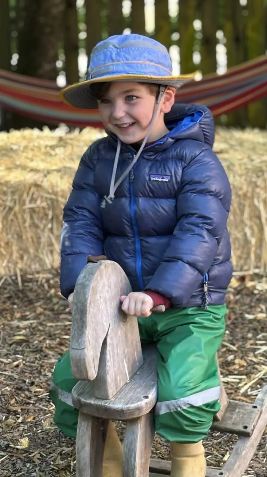 A preschooler in a sun hat riding a wooden rocking horse outdoors