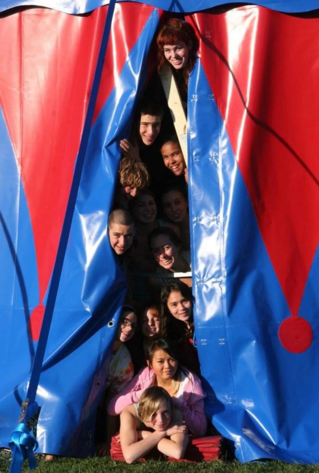 A group of campers peek smiling through the gap in the school's red-and-blue big-top circus tent, lit by afternoon sun