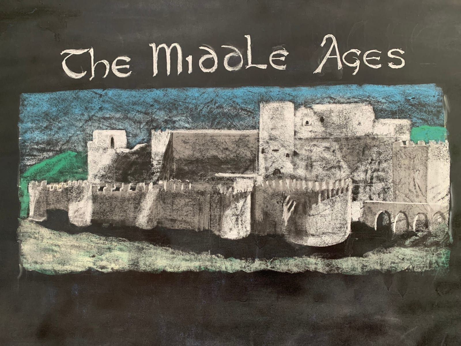 A chalk-pastel classroom blackboard drawing titled 'The Middle Ages,' showing a medieval walled castle with towers and a gatehouse, rendered in white, blue, and green chalk.