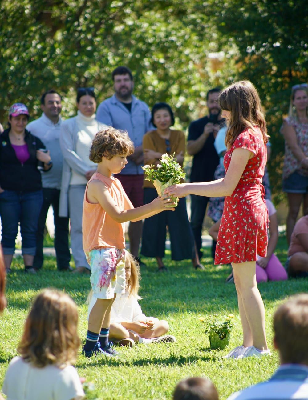 A boy in an orange tank top receives a small bouquet of yellow roses from an older student in a red floral dress, while younger children sit on the lawn nearby and a crowd of parents watches from behind
