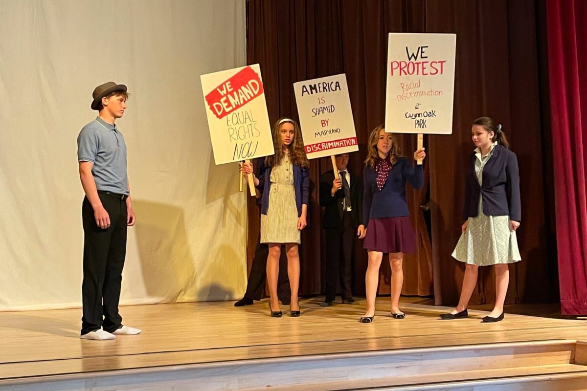 Four Summerfield High School students perform on an auditorium stage, four of them holding hand-lettered protest signs reading 'We Demand Equal Rights Now' and 'We Protest Racial Discrimination'.
