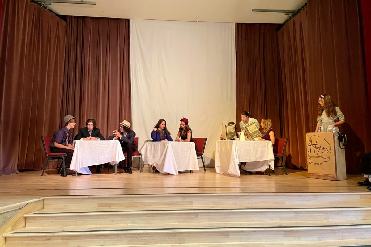 Students in period costume rehearse a diner scene on stage, seated at cloth-covered tables in front of a lectern labeled 'Hooper's', with the house lights up.