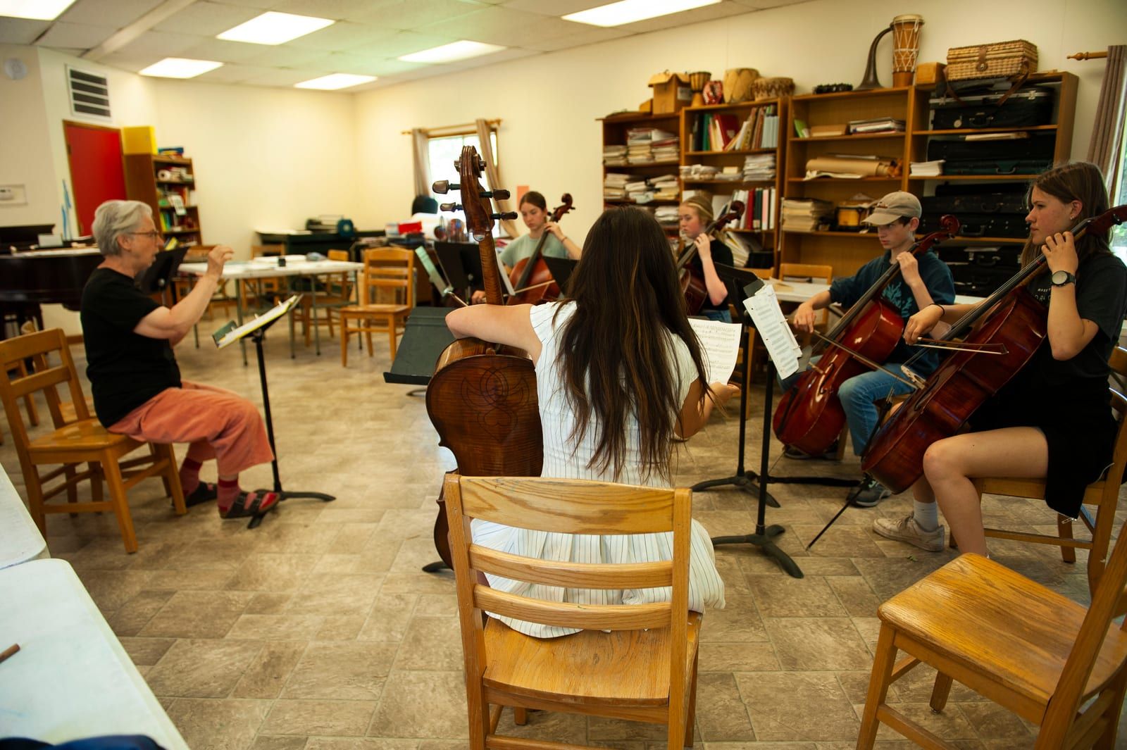 A small string ensemble of high school cello students plays together in a music room while a teacher conducts from a chair, sheet music stands and instrument cases around them.